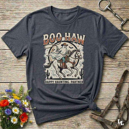 A dark heather gray graphic t-shirt laid flat on a rustic wooden surface. The shirt features a western-style illustration of a cowboy riding a horse with the text 'BOO HAW' above and 'HAPPY HAUNTING, PARTNER' below.