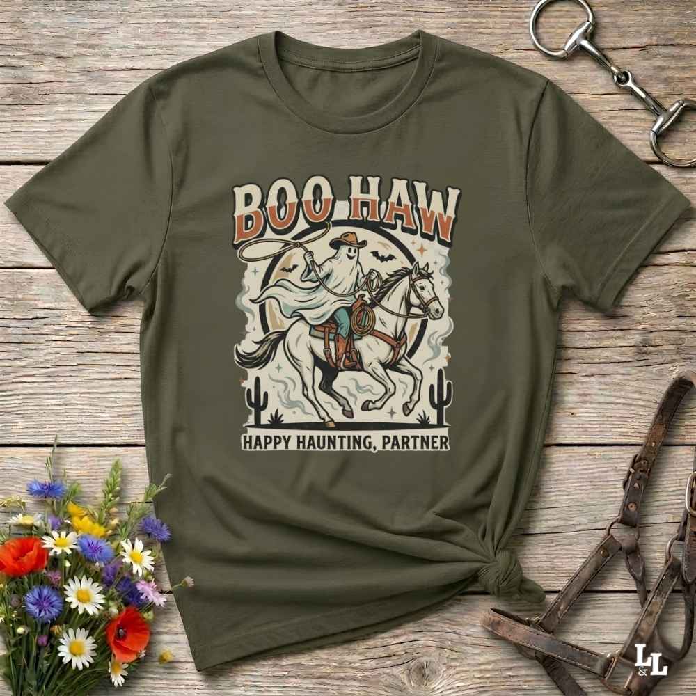 An olive green graphic t-shirt featuring a western-themed illustration of a cowboy on a horse with the text 'BOO HAW' and 'HAPPY HAUNTING, PARTNER'. The t-shirt is styled in a flat-lay on a rustic wooden background with decorative elements including a flower bouquet, leather halter, and horseshoe.