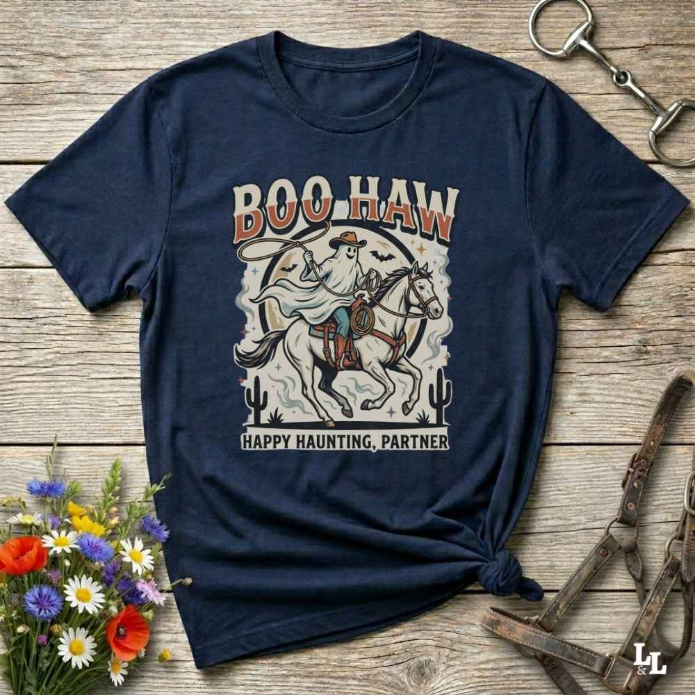 A navy blue graphic t-shirt laid flat on a rustic wooden surface, styled with equestrian and wildflower props. The shirt features a vintage-style cowboy on a horse with the text 'BOO HAW' and 'HAPPY HAUNTING, PARTNER'.