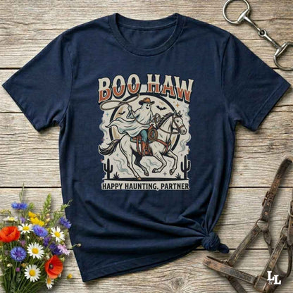 A navy blue graphic t-shirt laid flat on a rustic wooden surface, styled with equestrian and wildflower props. The shirt features a vintage-style cowboy on a horse with the text 'BOO HAW' and 'HAPPY HAUNTING, PARTNER'.