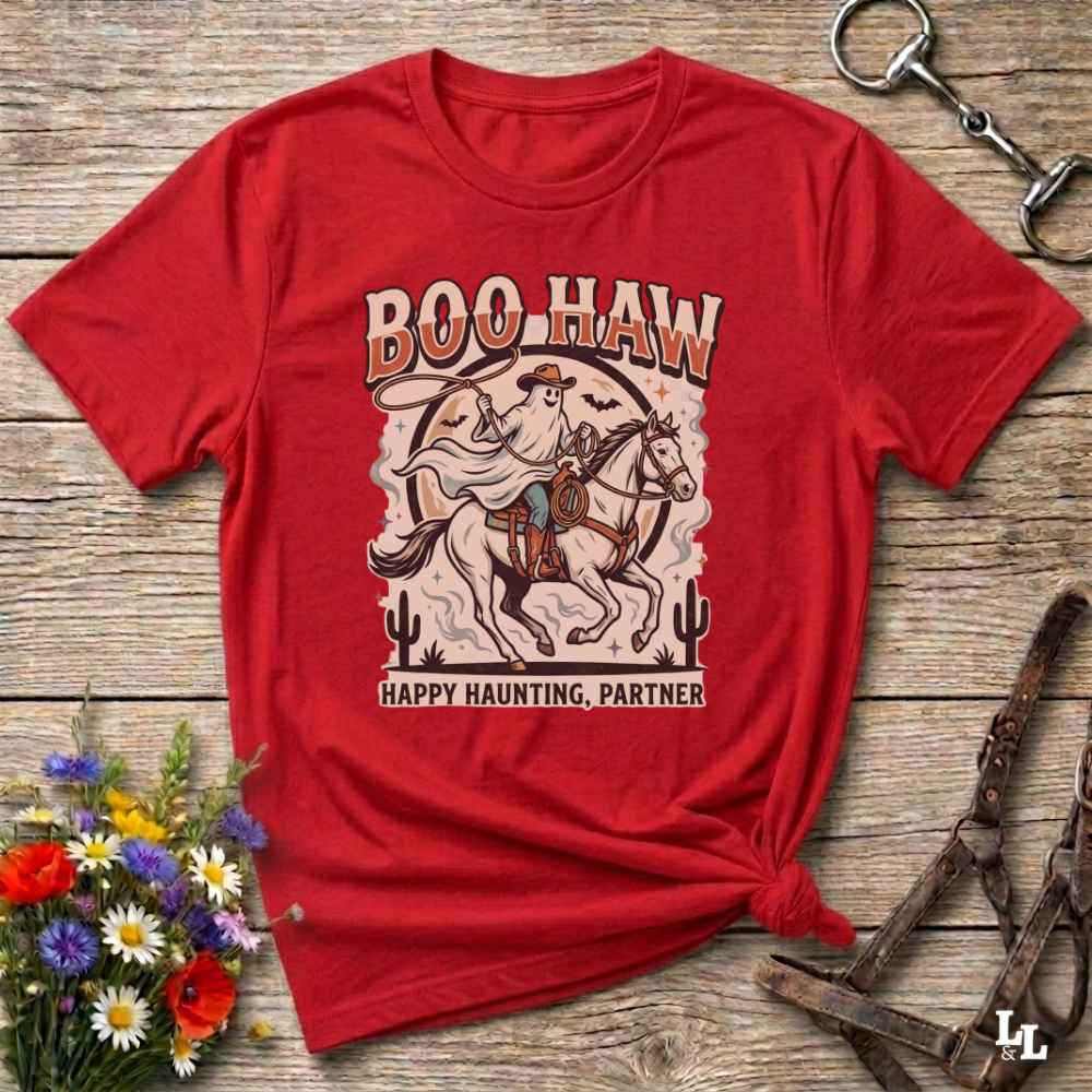 A red short-sleeve graphic T-shirt laid flat on a rustic wood surface, styled with flowers, a horse bit, and a leather bridle. The shirt has a large western-style ghostly rider on a horse graphic with the text 'BOO-HAW' and 'HAPPY HAUNTING, PARTNER'.