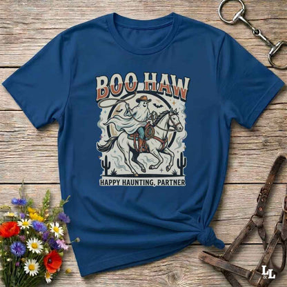 A navy blue graphic t-shirt with a western-themed 'Boo Haw Happy Haunting, Partner' design, styled in a flat-lay on a rustic wooden background with a horse halter, a small bouquet of wildflowers, and a metal tack strap.
