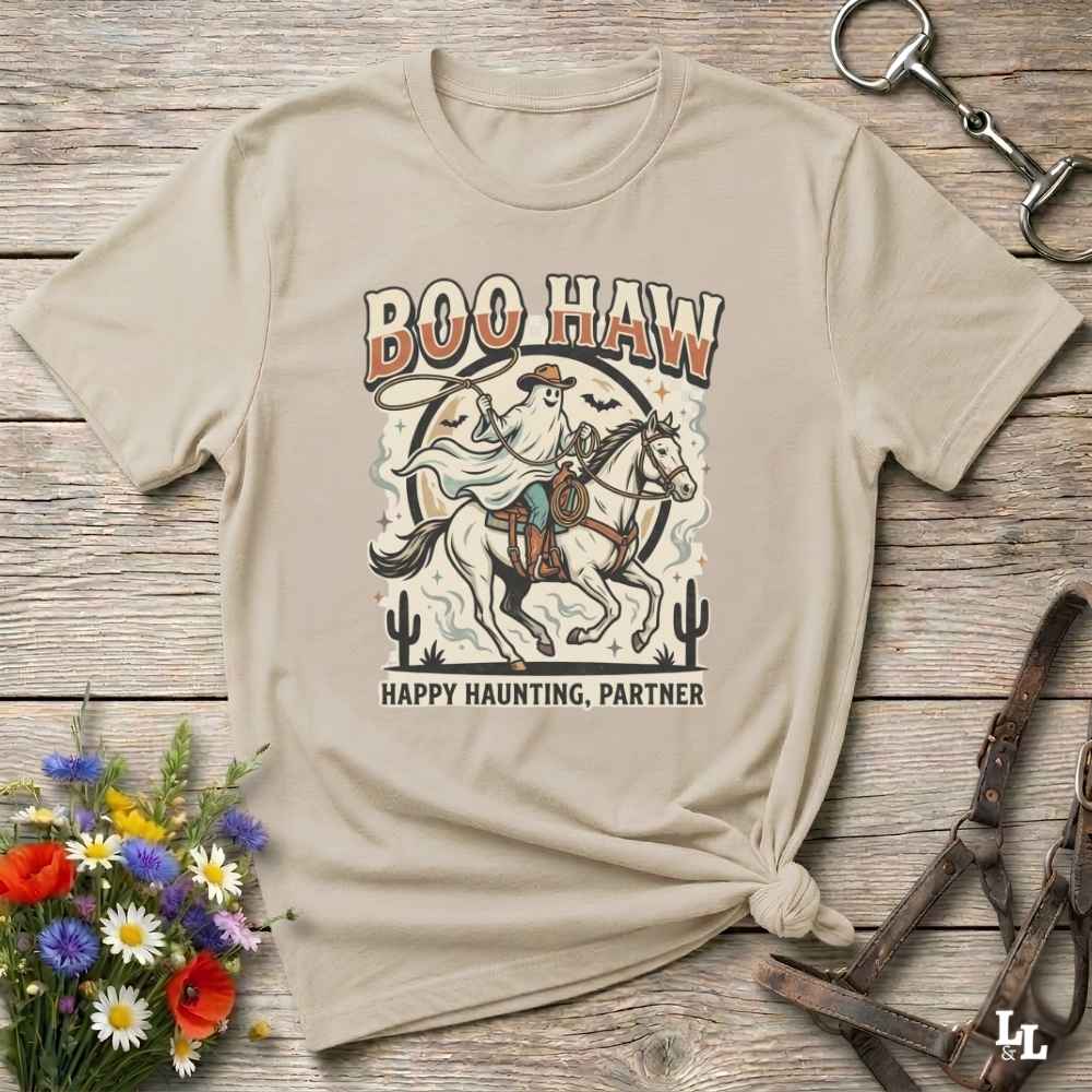 A beige graphic t-shirt with a western-themed 'Boo Haw Happy Haunting, Partner' design, styled flat on a rustic wooden background with decorative flowers and horse tack.