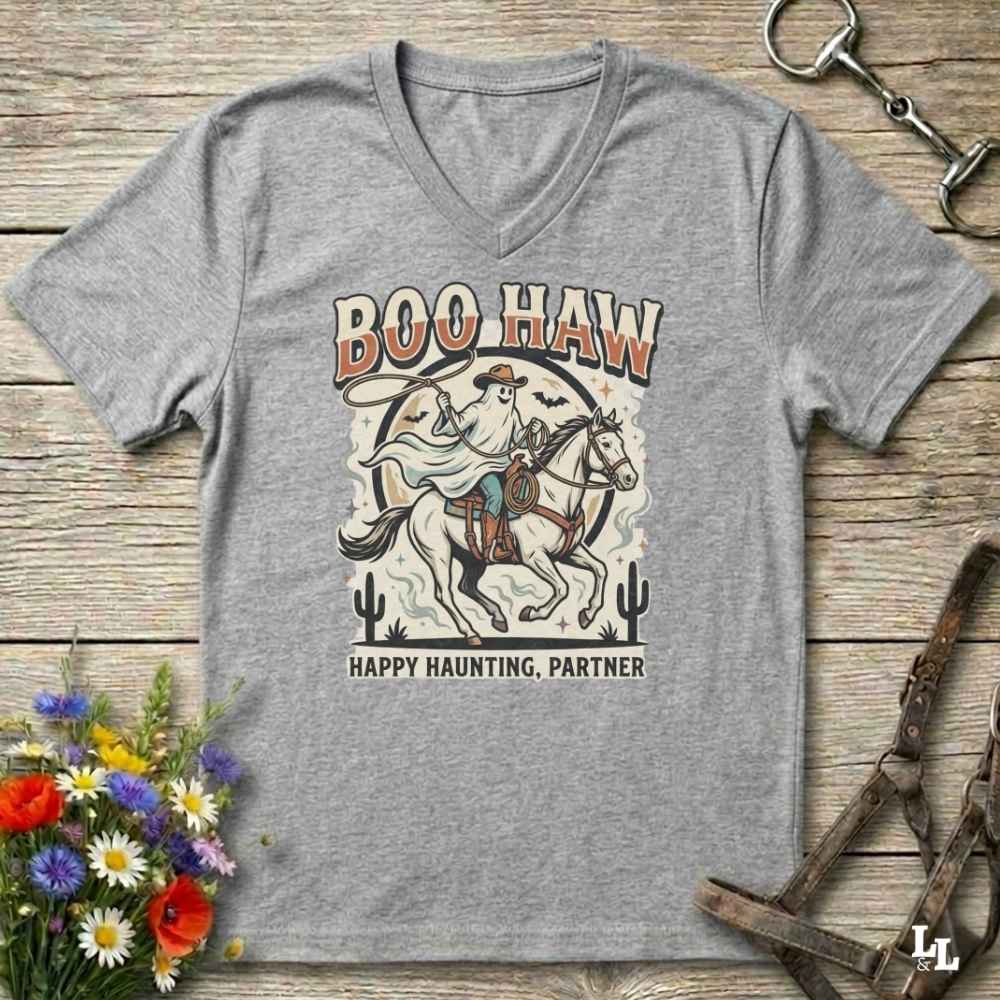 A heather gray V-neck t-shirt with a western-themed graphic print featuring a cowboy on a horse and the text 'BOO HAW' and 'HAPPY HAUNTING, PARTNER'. The shirt is displayed flat on a rustic wooden background with decorative horse tack and flowers.