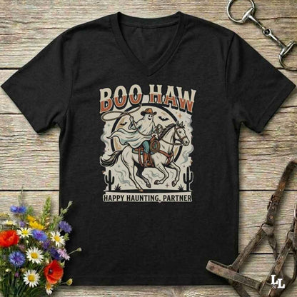 A black V-neck t-shirt with a western-themed graphic print reading 'BOO HAW' and 'HAPPY HAUNTING, PARTNER' around an illustration of a ghostly cowboy on a horse, displayed on a rustic wooden background with decorative flowers and horse tack.