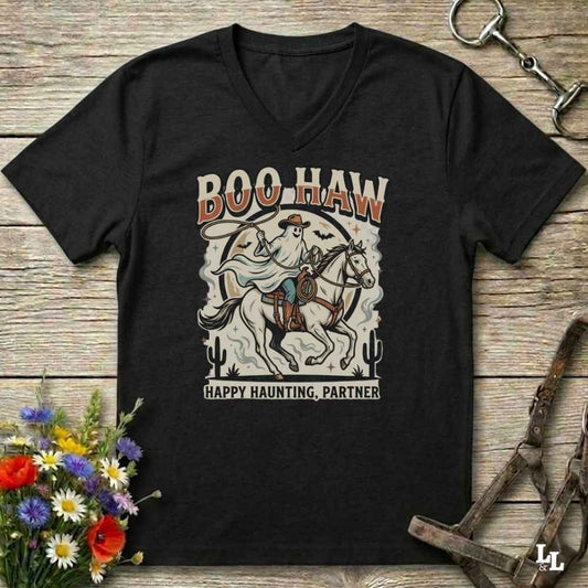 A black V-neck t-shirt with a western-themed graphic print reading 'BOO HAW' and 'HAPPY HAUNTING, PARTNER' around an illustration of a ghostly cowboy on a horse, displayed on a rustic wooden background with decorative flowers and horse tack.
