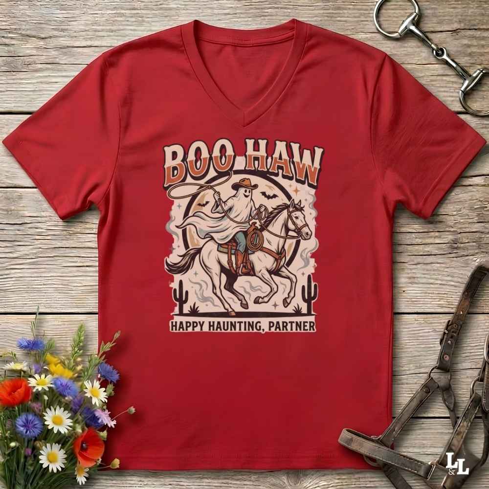 A red V-neck t-shirt with a western-themed 'Boo Haw Happy Haunting, Partner' graphic, laid flat on a rustic wooden background with equestrian and floral props.