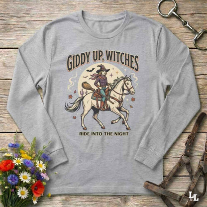 A light gray long-sleeve t-shirt with a vintage-style graphic of a witch riding a white horse, surrounded by the text 'GIDDY UP, WITCHES' and 'RIDE INTO THE NIGHT'. The shirt is displayed in a flat-lay arrangement on a rustic wooden background, styled with a bouquet of colorful wildflowers, a leather bridle, and a horse bit.