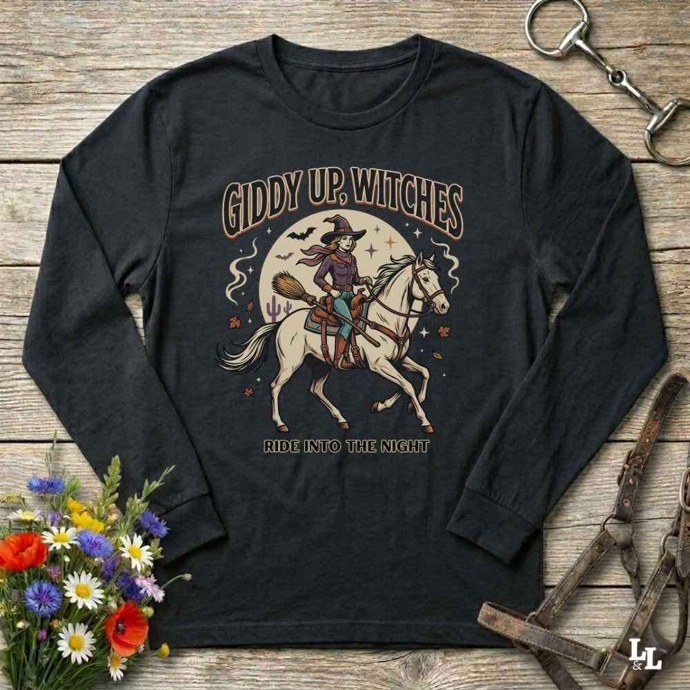 A black long-sleeve graphic t-shirt laid flat on a rustic wooden surface, surrounded by Halloween-themed props including a bouquet of wildflowers, a horse bridle, and a metal latch. The shirt features a graphic of a witch riding a white horse under a full moon with the text 'GIDDY UP, WITCHES' and 'RIDE INTO THE NIGHT'.
