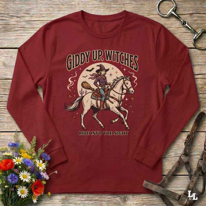 A long-sleeve crewneck t-shirt in maroon red, laid flat on a rustic wooden background. The shirt features a graphic of a witch riding a white horse into the night under a full moon, with the text 'GIDDY UP, WITCHES' above and 'RIDE INTO THE NIGHT' below.