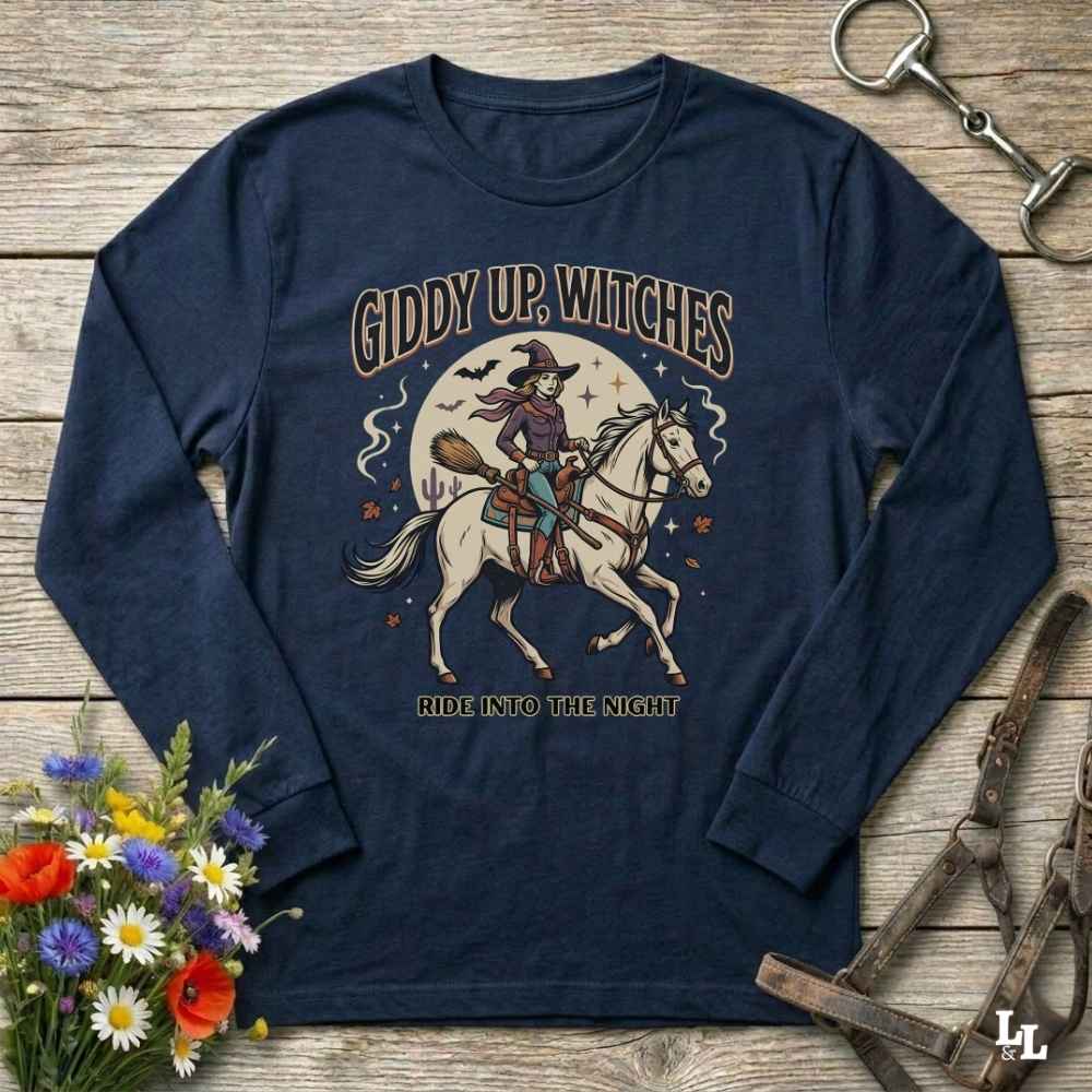 A navy blue long-sleeve t-shirt with a vintage-style graphic of a witch riding a white horse on a wooden background. The graphic features the text 'GIDDY UP, WITCHES' and 'RIDE INTO THE NIGHT' with an illustration of a witch on a broomstick and a moon.