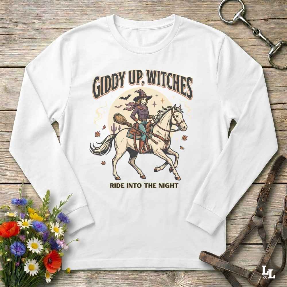 A white long-sleeve crewneck t-shirt with a vintage-style Halloween graphic, laid flat on a rustic wooden background with equestrian and floral props.