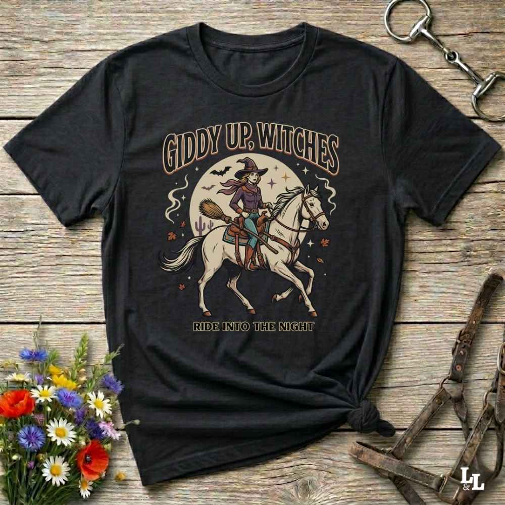 A black graphic t-shirt laid flat on a rustic wooden surface, styled with a small bouquet of flowers and equestrian tack. The shirt features a graphic of a witch riding a white horse under a full moon with the text 'GIDDY UP, WITCHES' and 'RIDE INTO THE NIGHT'.