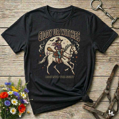 A black graphic t-shirt laid flat on a rustic wooden surface, styled with a small bouquet of flowers and equestrian tack. The shirt features a graphic of a witch riding a white horse under a full moon with the text 'GIDDY UP, WITCHES' and 'RIDE INTO THE NIGHT'.