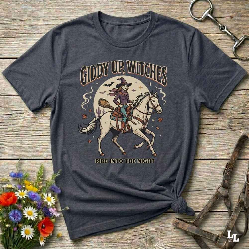 A dark heather gray graphic t-shirt laid flat on a rustic wooden surface. The shirt features a vintage-style illustration of a witch riding a white horse under a full moon, with the text 'GIDDY UP, WITCHES' and 'RIDE INTO THE NIGHT'.