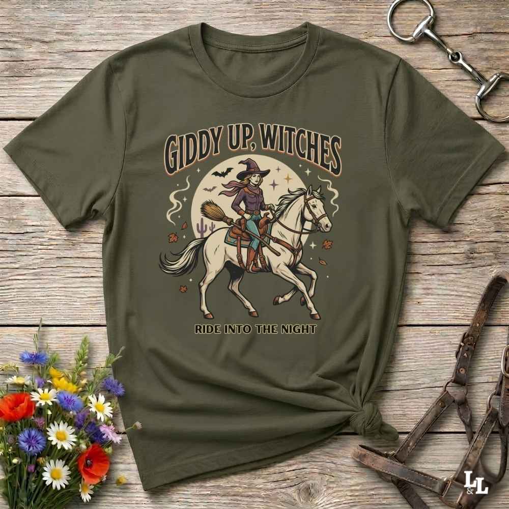An olive green graphic t-shirt laid flat on a rustic wooden surface, styled with a small bouquet of flowers, a horse bit, and leather tack. The t-shirt features a graphic of a witch riding a white horse under a full moon with the text 'GIDDY UP, WITCHES' and 'RIDE INTO THE NIGHT'.