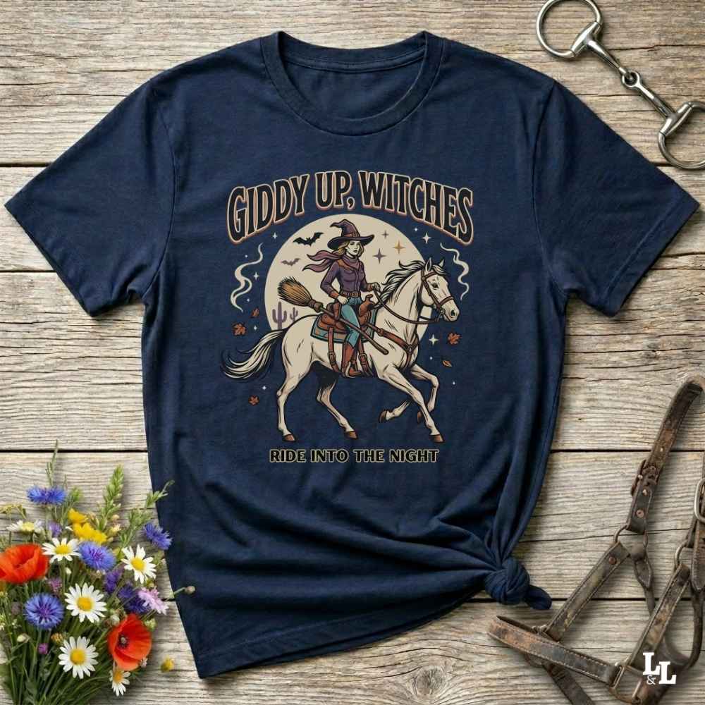 A navy blue graphic t-shirt featuring a vintage-style illustration of a witch riding a white horse under a full moon, with the text 'GIDDY UP, WITCHES' and 'RIDE INTO THE NIGHT'. The t-shirt is styled in a flat-lay on a rustic wooden background with decorative flowers and horse tack.