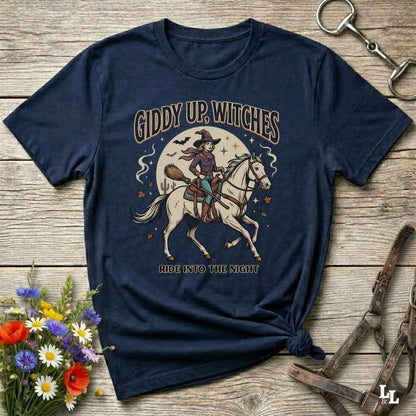 A navy blue graphic t-shirt featuring a vintage-style illustration of a witch riding a white horse under a full moon, with the text 'GIDDY UP, WITCHES' and 'RIDE INTO THE NIGHT'. The t-shirt is styled in a flat-lay on a rustic wooden background with decorative flowers and horse tack.