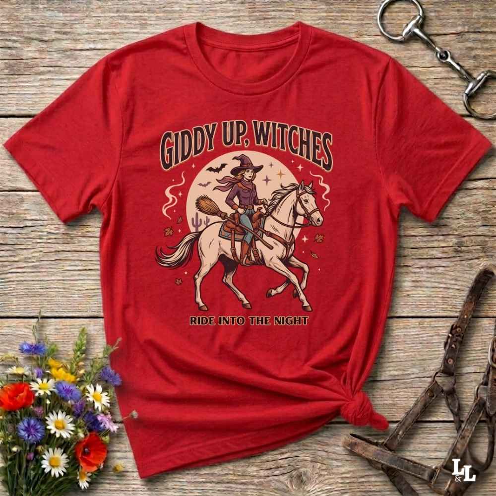 A red short-sleeve graphic T-shirt laid flat on a rustic wooden surface, styled with flowers, a horse bit, and a leather bridle. The shirt features a large front print of a witch riding a white horse under the text 'GIDDY UP, WITCHES' and 'RIDE INTO THE NIGHT'.