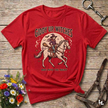 A red short-sleeve graphic T-shirt laid flat on a rustic wooden surface, styled with flowers, a horse bit, and a leather bridle. The shirt features a large front print of a witch riding a white horse under the text 'GIDDY UP, WITCHES' and 'RIDE INTO THE NIGHT'.