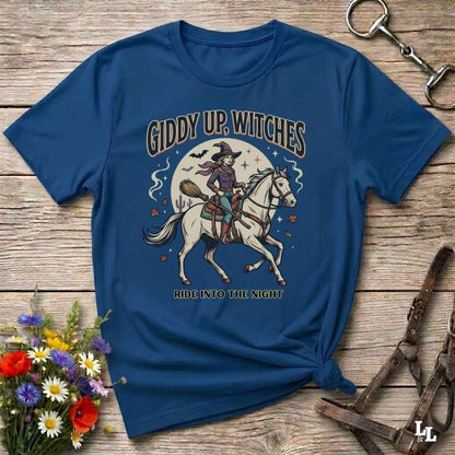 A blue graphic t-shirt featuring a witch riding a white horse under a full moon with the text 'GIDDY UP, WITCHES' and 'RIDE INTO THE NIGHT'. The shirt is styled in a flat-lay on a rustic wooden background with decorative flowers and horse tack.
