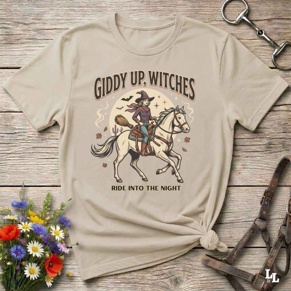 A beige graphic t-shirt with a Halloween-themed design of a witch riding a white horse, styled in a flat-lay on a rustic wooden background with flowers and horse tack.
