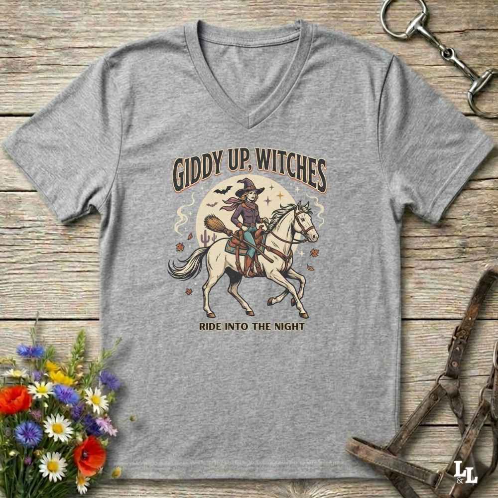 A heather gray V-neck t-shirt with a vintage-style Halloween graphic of a witch riding a white horse under the text 'GIDDY UP, WITCHES' and 'RIDE INTO THE NIGHT'. The shirt is displayed flat on a rustic wooden background with equestrian and floral props.