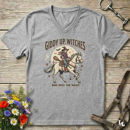 A heather gray V-neck t-shirt with a vintage-style Halloween graphic of a witch riding a white horse under the text 'GIDDY UP, WITCHES' and 'RIDE INTO THE NIGHT'. The shirt is displayed flat on a rustic wooden background with equestrian and floral props.