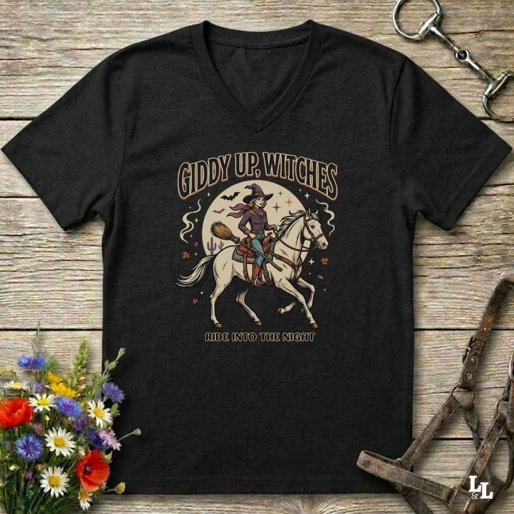 A black V-neck t-shirt with a vintage-style Halloween graphic of a witch riding a white horse under a full moon, with the text 'GIDDY UP, WITCHES' and 'ALL INTO THE NIGHT'. The shirt is displayed flat on a rustic wooden background with decorative horse tack and flowers.