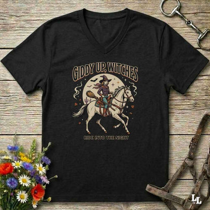 A black V-neck t-shirt with a vintage-style Halloween graphic of a witch riding a white horse under a full moon, with the text 'GIDDY UP, WITCHES' and 'ALL INTO THE NIGHT'. The shirt is displayed flat on a rustic wooden background with decorative horse tack and flowers.