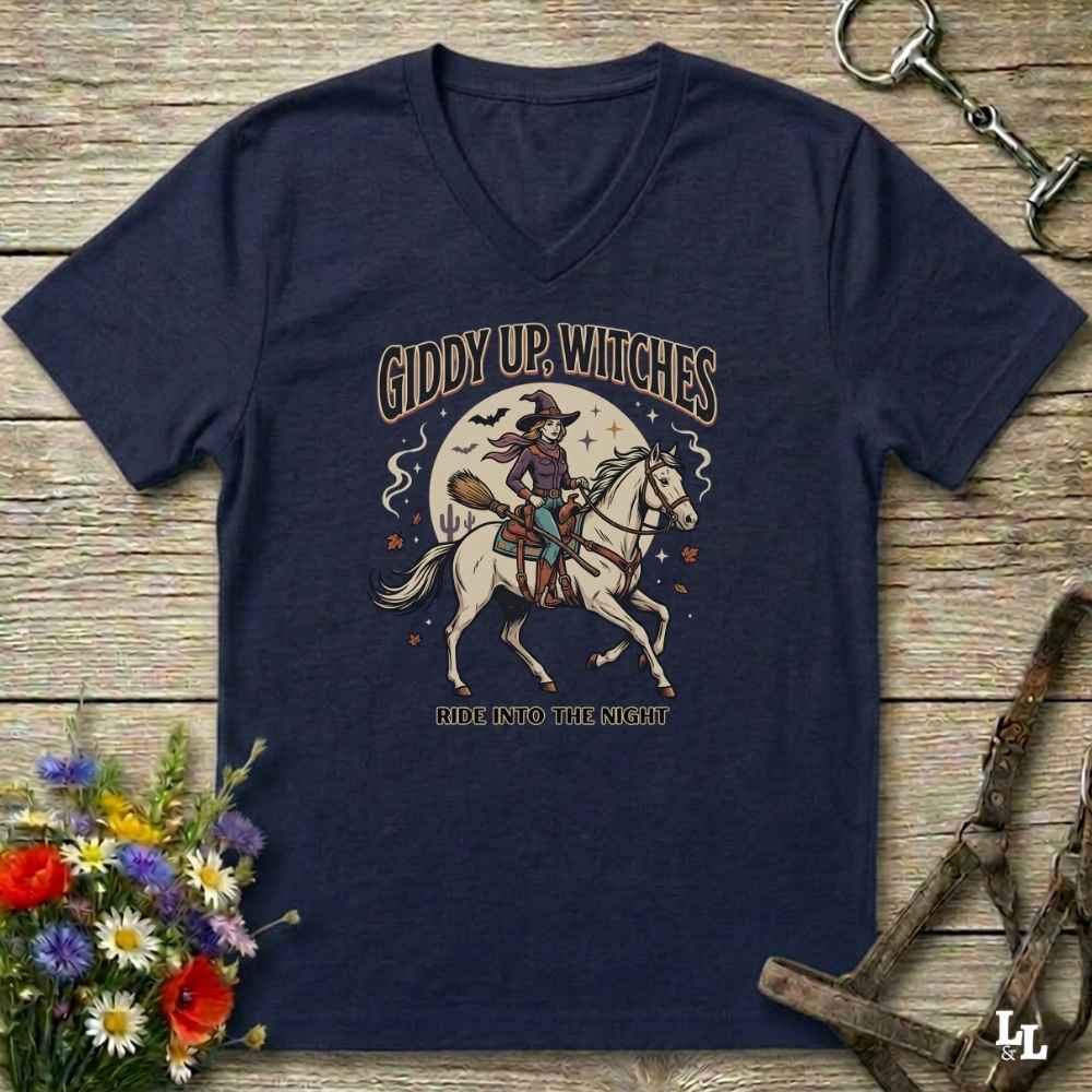 A navy blue V-neck t-shirt with a vintage-style Halloween graphic of a witch riding a white horse under a full moon, with the text 'GIDDY UP, WITCHES' and 'ALL INTO THE NIGHT'. The shirt is displayed flat on a rustic wooden background with decorative props including a bouquet of flowers and horse tack.