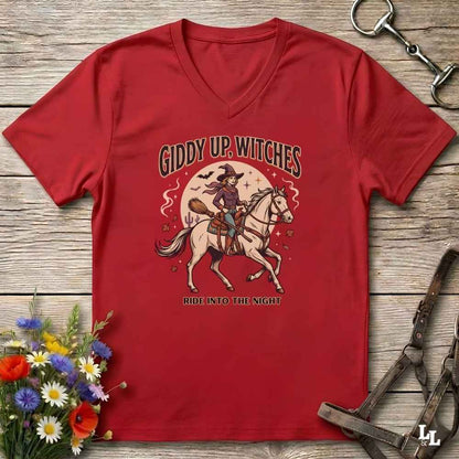 A red V-neck t-shirt with a vintage-style Halloween graphic of a witch riding a white horse under a full moon, with the text 'GIDDY UP, WITCHES' and 'RIDE INTO THE NIGHT'. The shirt is displayed flat on a rustic wooden background with decorative horse tack and flowers.