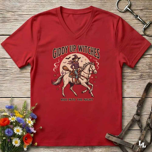A red V-neck t-shirt with a vintage-style Halloween graphic of a witch riding a white horse under a full moon, with the text 'GIDDY UP, WITCHES' and 'RIDE INTO THE NIGHT'. The shirt is displayed flat on a rustic wooden background with decorative horse tack and flowers.