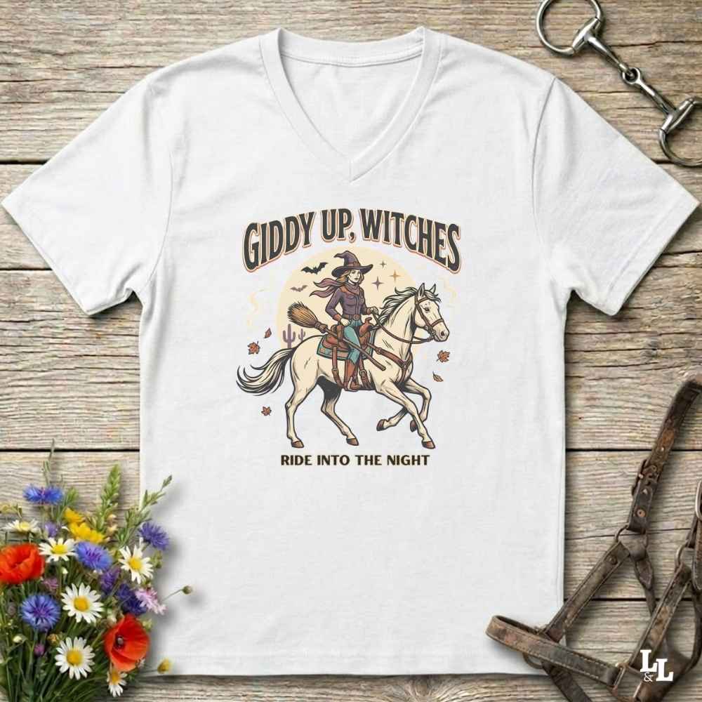 A white v-neck t-shirt with a graphic of a witch riding a white horse under the text 'GIDDY UP, WITCHES' and 'RIDE INTO THE NIGHT', displayed on a rustic wooden background with decorative flowers and equestrian accessories.