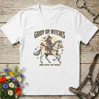 A white v-neck t-shirt with a graphic of a witch riding a white horse under the text 'GIDDY UP, WITCHES' and 'RIDE INTO THE NIGHT', displayed on a rustic wooden background with decorative flowers and equestrian accessories.
