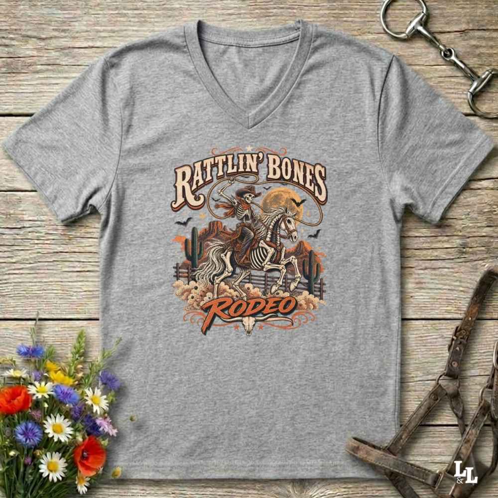 A heather gray Halloween V-neck t-shirt with a detailed western skeleton cowboy graphic on the chest, styled in a flat-lay on a rustic wooden background with equestrian and floral props.