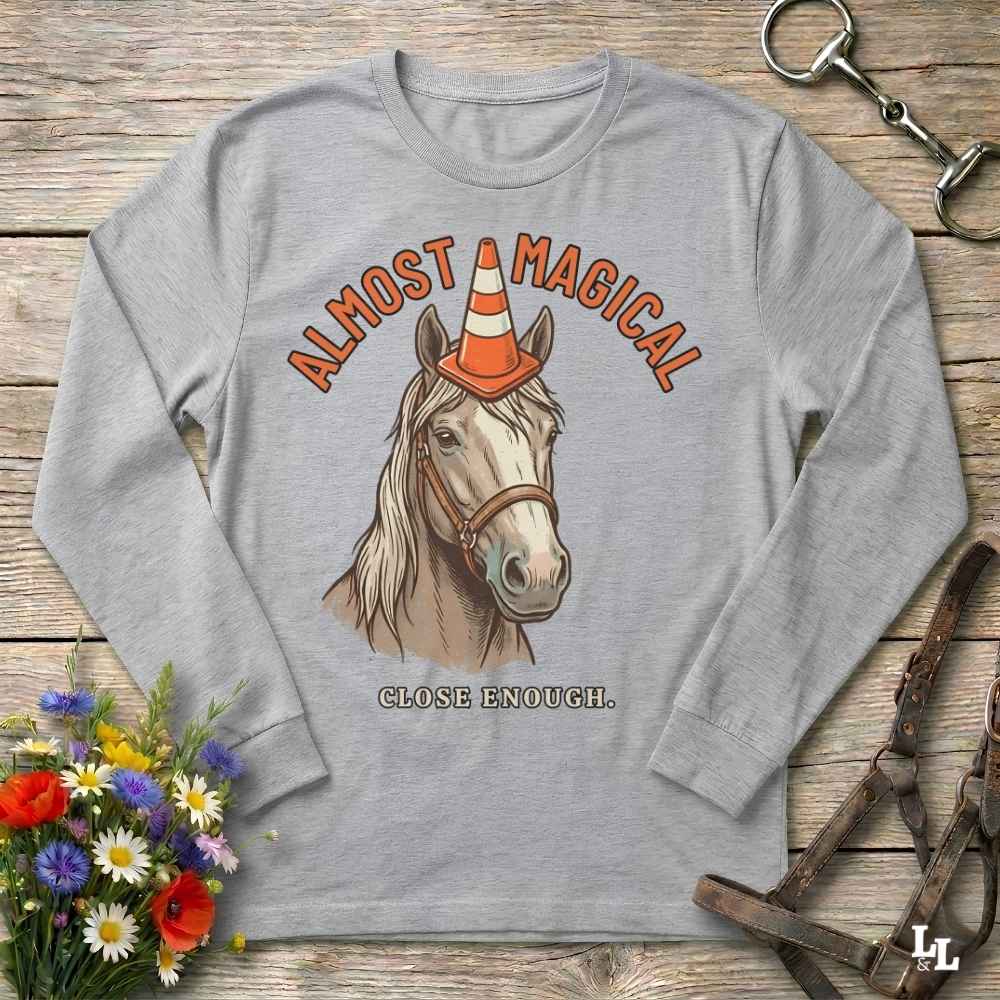 A light gray long-sleeve t-shirt with a graphic of a horse wearing an orange and white traffic cone horn, with the text 'ALMOST MAGICAL' above and 'CLOSE ENOUGH.' below, laid flat on a rustic wooden surface with decorative flowers and leather tack around it.
