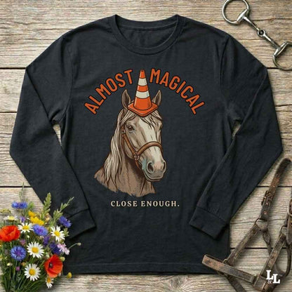 A black long-sleeve graphic t-shirt laid flat on a rustic wooden surface, featuring an illustration of a horse wearing an orange and white traffic cone hat, with the text 'ALMOST MAGICAL' above and 'CLOSE ENOUGH.' below.