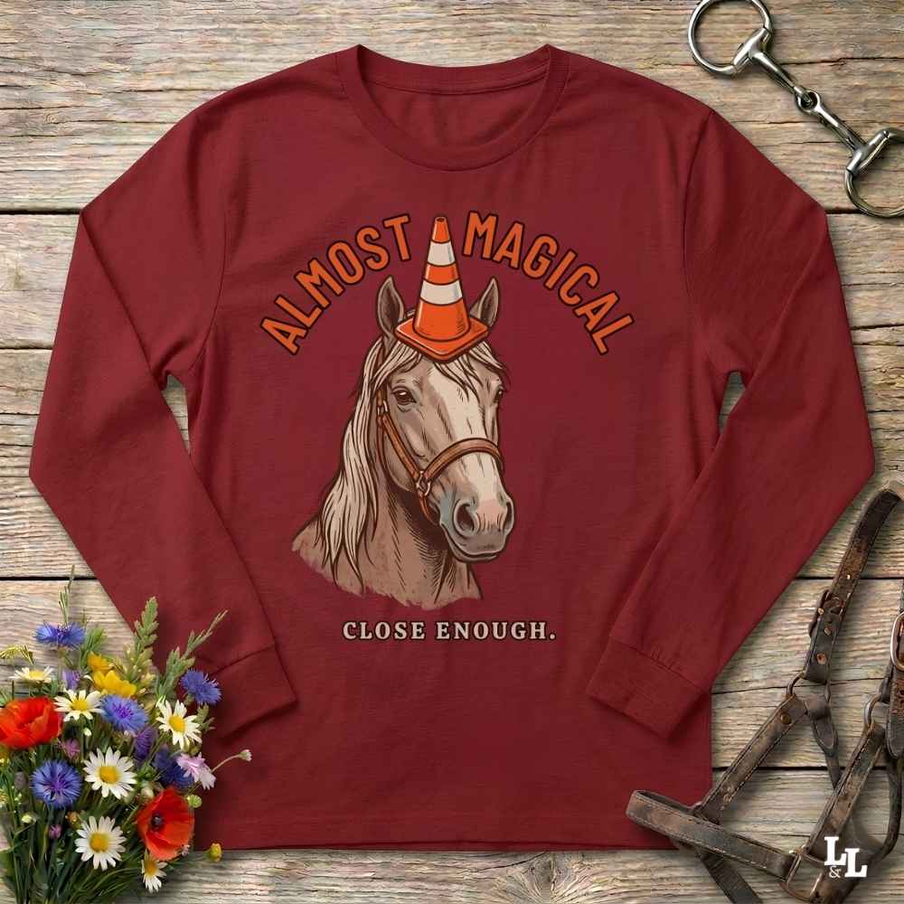 A long-sleeve crewneck shirt in deep burgundy, featuring a graphic of a horse wearing an orange and white traffic cone with the text 'ALMOST MAGICAL' above and 'CLOSE ENOUGH.' below.