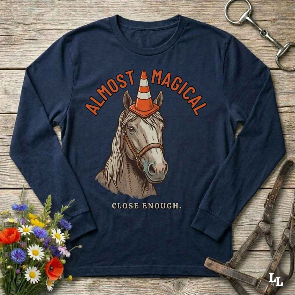 A navy blue long-sleeve graphic t-shirt featuring an illustration of a horse wearing an orange and white traffic cone, with the text 'ALMOST MAGICAL' above and 'CLOSE ENOUGH.' below.