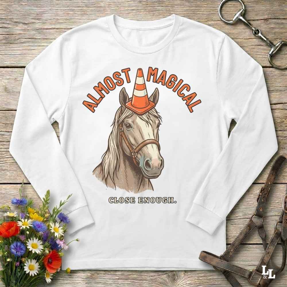 A white long-sleeve t-shirt with a graphic of a horse wearing an orange and white traffic cone hat, with the text 'ALMOST MAGICAL' above and 'CLOSE ENOUGH.' below, displayed on a rustic wooden background with equestrian accessories and flowers.