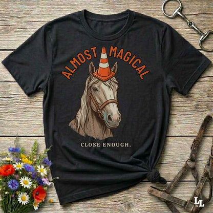 A black graphic t-shirt laid flat on a rustic wooden surface, featuring an illustration of a horse wearing an orange and white traffic cone hat, with the text 'ALMOST MAGICAL' above and 'CLOSE ENOUGH.' below.