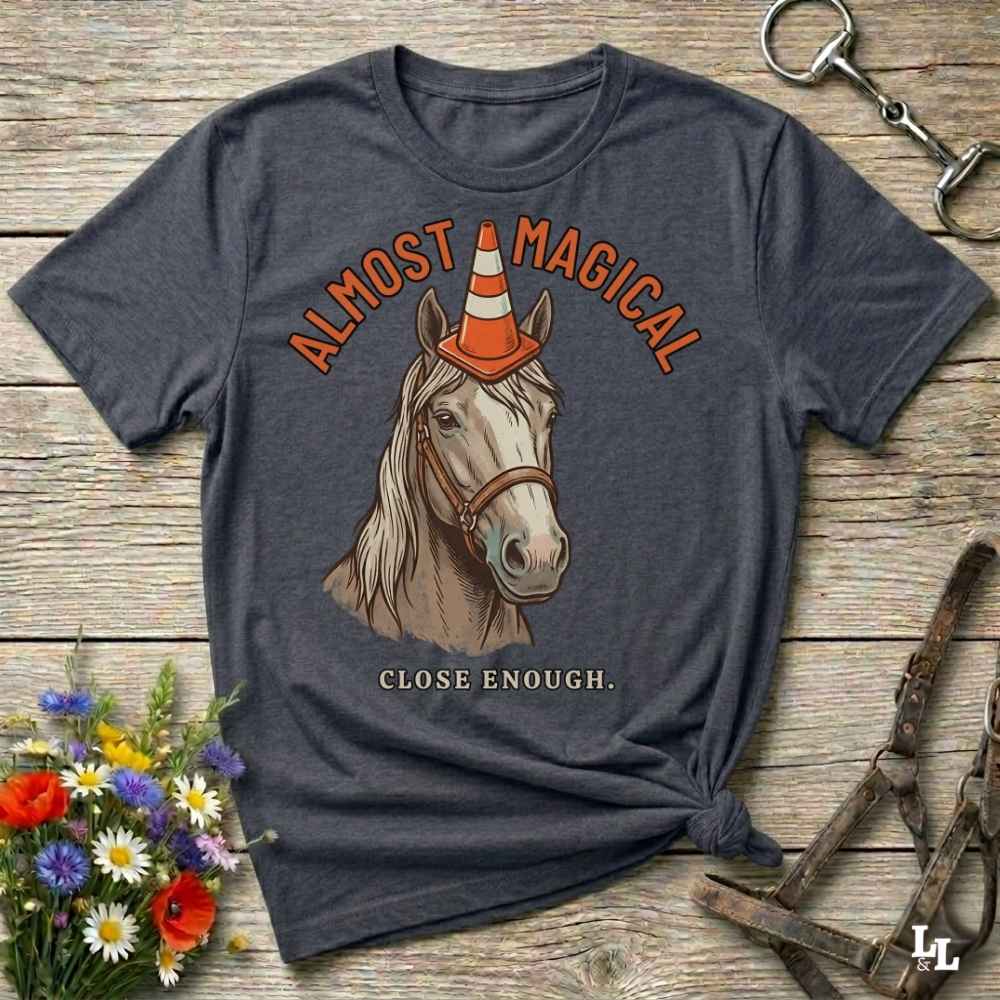 A dark charcoal gray graphic t-shirt laid flat on a rustic wooden surface, styled with equestrian props like a halter, a horse bit, and a small bouquet of wildflowers. The t-shirt features a graphic of a white horse wearing an orange and white traffic cone as a horn, with the text 'ALMOST MAGICAL' above and 'CLOSE ENOUGH.