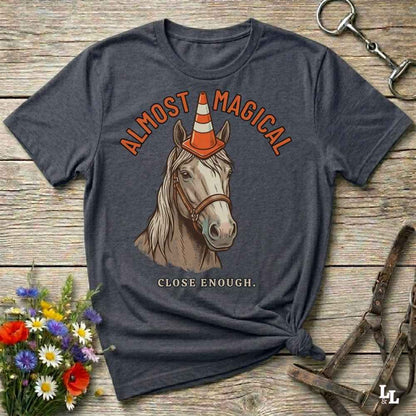 A dark charcoal gray graphic t-shirt laid flat on a rustic wooden surface, styled with equestrian props like a halter, a horse bit, and a small bouquet of wildflowers. The t-shirt features a graphic of a white horse wearing an orange and white traffic cone as a horn, with the text 'ALMOST MAGICAL' above and 'CLOSE ENOUGH.