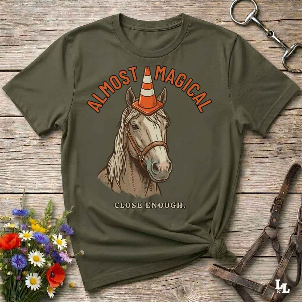 An olive green graphic t-shirt laid flat on a rustic wooden surface, styled with equestrian accessories like a horse bit, leather bridle, and a small bouquet of wildflowers. The t-shirt features a graphic of a white horse wearing an orange and white traffic cone as a hat, with the text 'ALMOST MAGICAL' above and 'CLOSE ENOUGH.