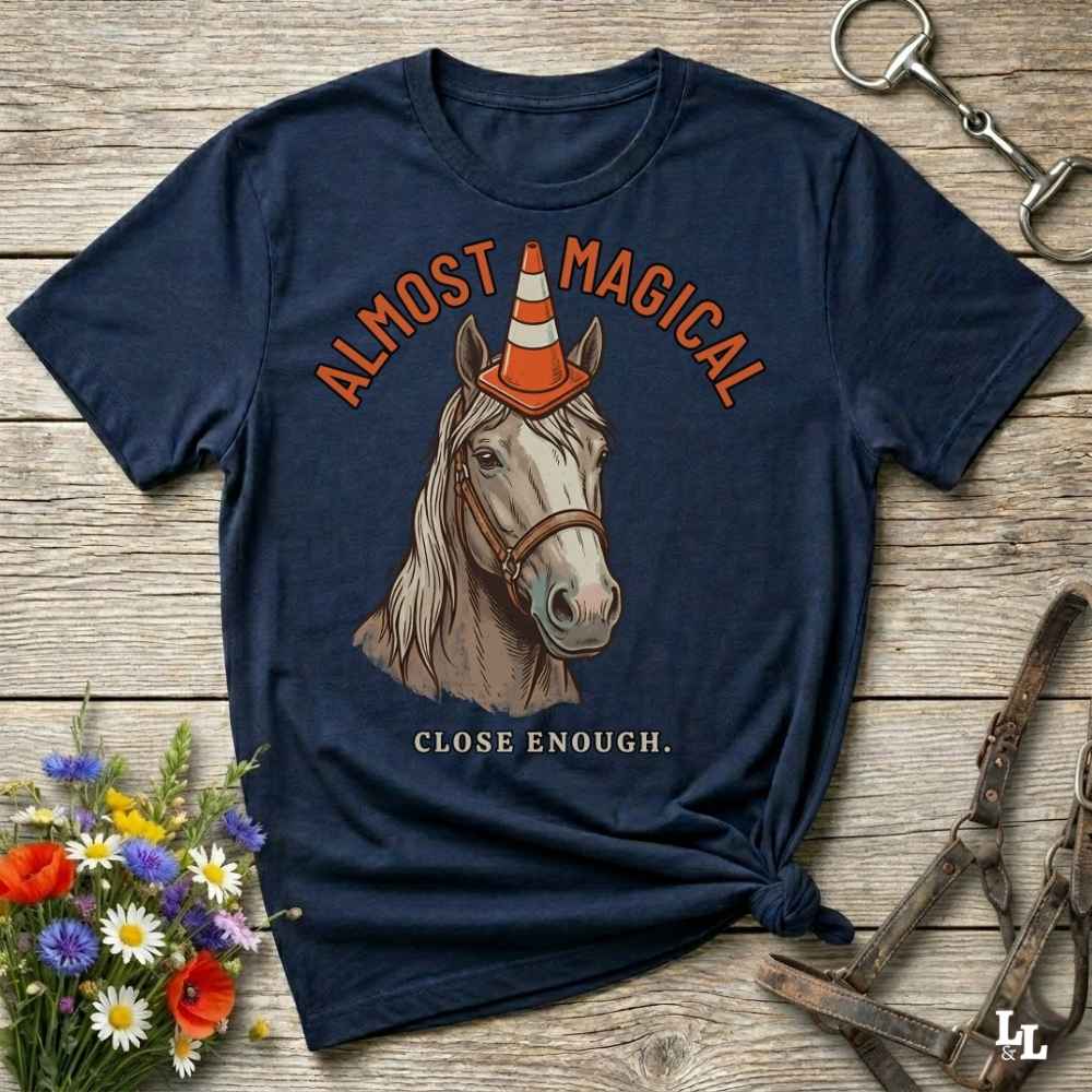 A navy blue graphic t-shirt laid flat on a rustic wooden surface, styled with equestrian accessories and a small bouquet of wildflowers. The shirt features a graphic of a white horse wearing a traffic cone hat, with the text 'ALMOST MAGICAL' above and 'CLOSE ENOUGH.
