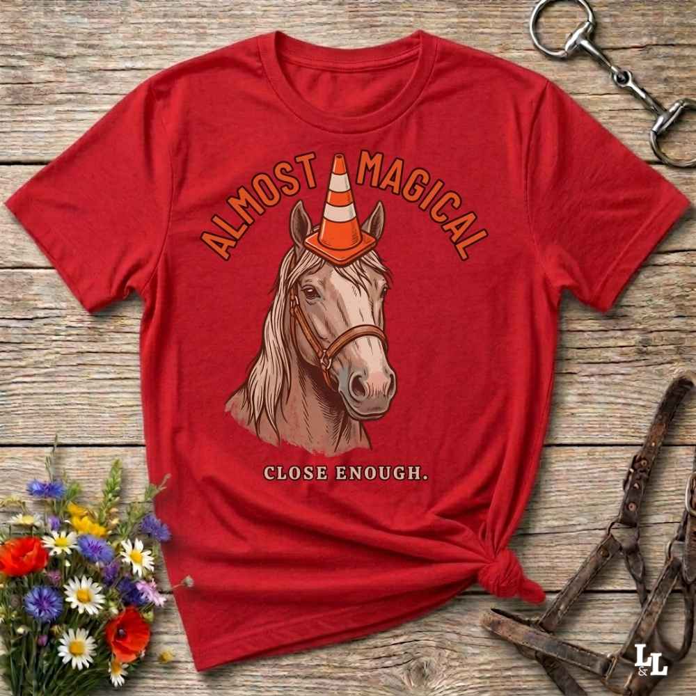 A red graphic t-shirt featuring an illustration of a horse wearing an orange and white traffic cone, with the text 'ALMOST MAGICAL' above and 'CLOSE ENOUGH.' below.