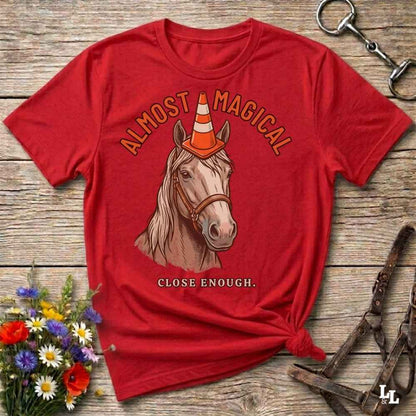 A red graphic t-shirt featuring an illustration of a horse wearing an orange and white traffic cone, with the text 'ALMOST MAGICAL' above and 'CLOSE ENOUGH.' below.