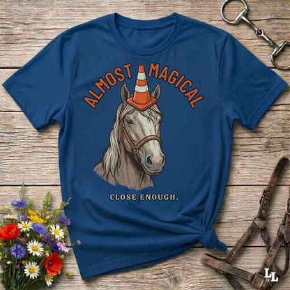 A navy blue graphic t-shirt laid flat on a rustic wooden surface, styled with equestrian accessories and a small bouquet of wildflowers. The shirt features a graphic of a white horse wearing an orange and white traffic cone, with the text 'ALMOST MAGICAL' above and 'CLOSE ENOUGH.