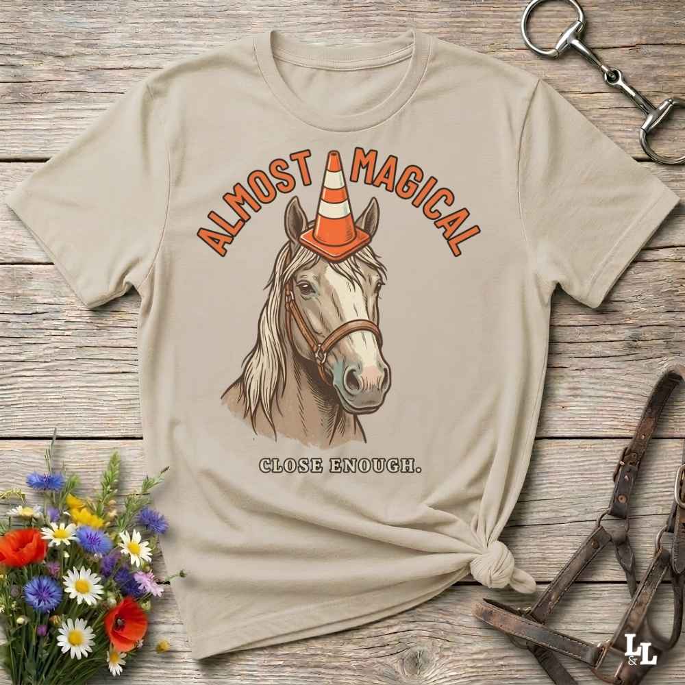 A beige graphic t-shirt featuring an illustration of a horse wearing an orange traffic cone as a hat, with the text 'ALMOST MAGICAL' above and 'CLOSE ENOUGH.' below.
